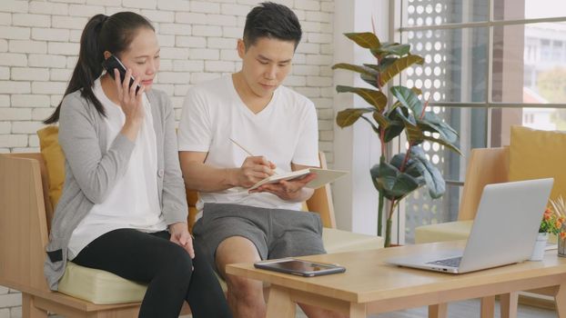 Happy Asian family couple husband and wife working confirming booking having talking call phone conversation and making writing notes of information together from home in living room. small business