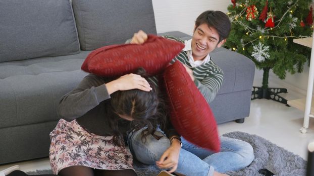 Happy Asian beautiful young family couple husband and wife smile and laugh enjoying funny fighting with pillows in living room with winter day at home.
