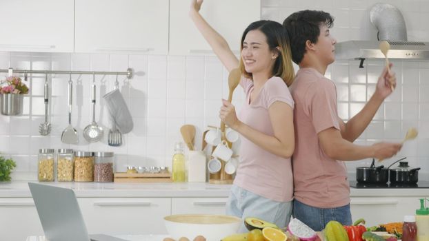 Happy Asian beautiful family couple husband and wife singing in kitchenware microphones in kitchen together having fun dance listen music at home. Two people dancing