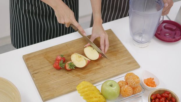 Happy Asian beautiful young family couple husband and wife making fresh smoothie in kitchen together at home. The man and woman slicing fresh apple fruits and veggies. Healthy lifestyle concept