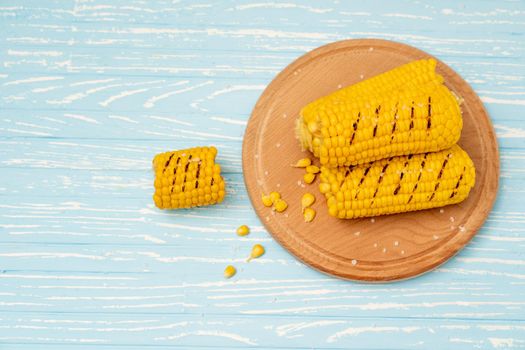 Grilled hot Corn cob lies on round cutting board plate blue wooden table background. Copy space for text.