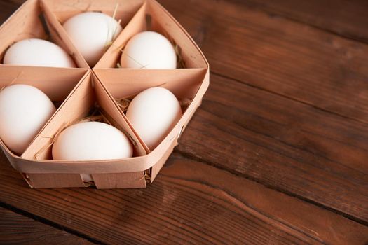 White chicken eggs lie in round wooden basket which stands on a dark wooden table. Easter background. Seasonal holiday flat lay with free space for text.