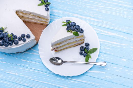 Appetizing cheesecake biscuit pillow decorated white cream blueberries and mint stands on wooden blue rustic table. Sweet cake with piece and spoon on plate