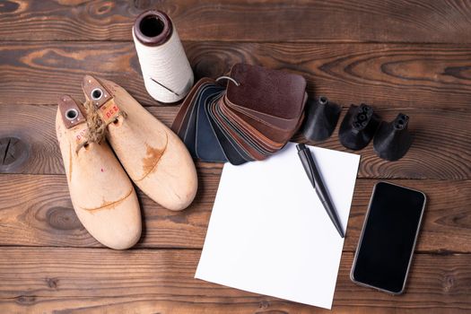 Leather samples for shoes and wooden shoe last on dark wooden table with Empty white sheet of paper for notes.. Designer furniture clothes. Shoe maker workspace.