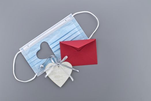Medical mask with cut out heart shape, red rose and wooden heart medallion on gray background with free copy space