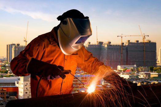 Industrial worker welding steel structure for infrastructure building project with construction site of high rise building in background.