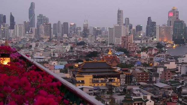 View of traditional and modern buildings of oriental city. Beautiful flowerbed against cityscape of traditional houses and skyscrapers on misty day on streets of Bangkok or Krungtep