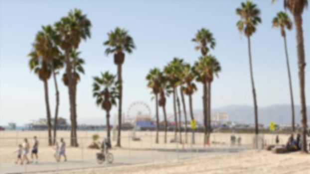 California beach aesthetic, people ride cycles on a bicycle path. Blurred, defocused background. Amusement park on pier and palms in Santa Monica american pacific ocean resort, Los Angeles CA USA.