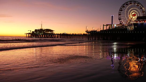 Twilight waves against classic illuminated ferris wheel, amusement park on pier in Santa Monica pacific ocean beach resort. Summertime iconic symbol of California glowing in dusk, Los Angeles, CA USA