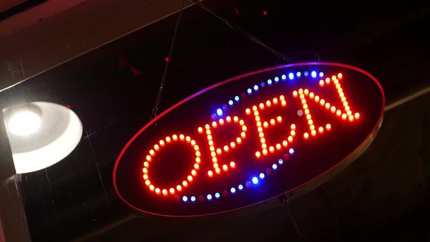 Open neon sign glowing in the dark. Vivid retro styled text at entrance on glass window. Colorful electric banner selective focus close up. Light bulbs radiance at night. Shiny illuminated lettering.