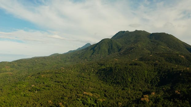 Mountains covered rainforest, trees and blue sky with clouds, aerial view. Camiguin, Philippines. Mountain landscape on tropical island with mountain peaks covered with forest. Slopes of mountains with evergreen vegetation.