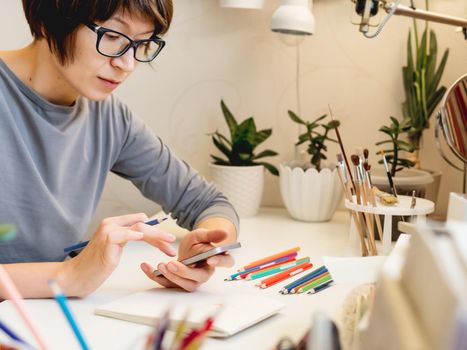 Woman with short hair cut is drawing in notebook with smartphone. Calming hobby, antistress leisure. Artist at work. Cozy workplace.