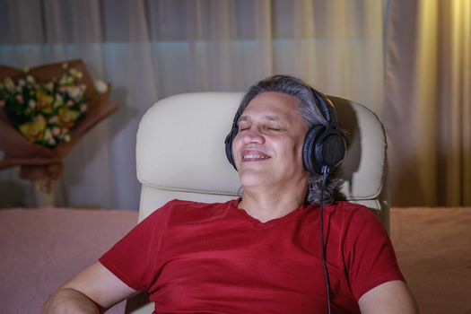 50-year-old man listens to music on headphones at home, sitting in a chair. Relaxing delight.