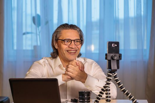 A 50-year-old blogger with glasses is recording an online seminar on his smartphone for his subscribers. With the help of modern devices, a computer and a smartphone, the master class is broadcast live.