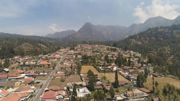 village in mountain valley. mountains with farmlands, village, fields with crops, trees. Aerial view farm lands on mountainside. tropical landscape Java Indonesia.
