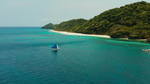 Sailing yacht in crystal clear turquoise water right next to the white sand beach of tropical island, aerial view. Sailing boat glides over the waves, Boracay, Philippines.