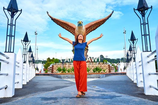 Happy girl tourist posing next to sculpture of a red eagle spreading its wings. Popular tourist spot on Langkawi island.