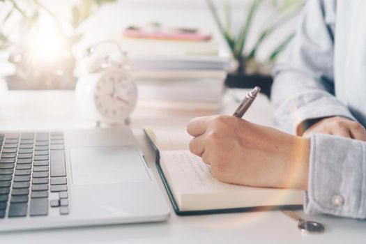 Woman is writing in planner notebook while using laptop to work or plan with workspace at home. Education business lifestyle concept.