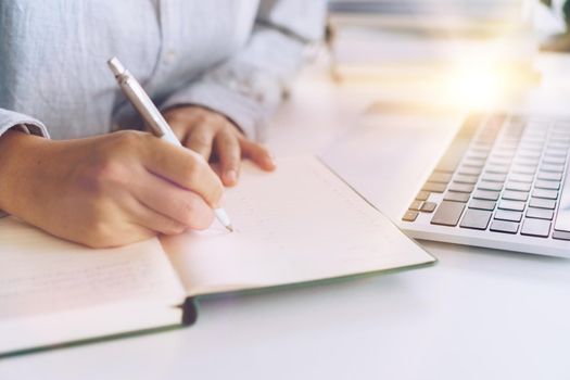 Woman is writing in planner notebook while using laptop to work or plan with workspace at home. Education business lifestyle concept.