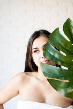 Spa Facial Mask. Spa and beauty. Happy beautiful brunette woman wearing bath towels holding a green monstera leaf in front of her face