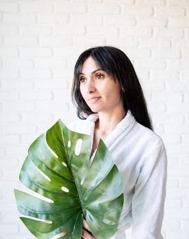 Spa Facial Mask. Spa and beauty. Happy beautiful brunette middle eastern woman wearing bath robes holding a green monstera leaf