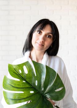 Spa Facial Mask. Spa and beauty. Happy beautiful brunette middle eastern woman wearing bath robes holding a green monstera leaf