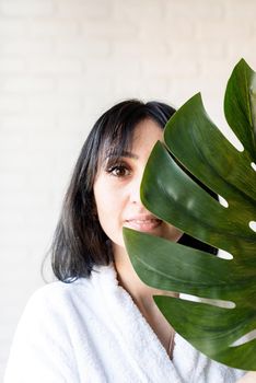 Spa Facial Mask. Spa and beauty. Happy beautiful brunette middle eastern woman wearing bath robes holding a green monstera leaf in front of her face