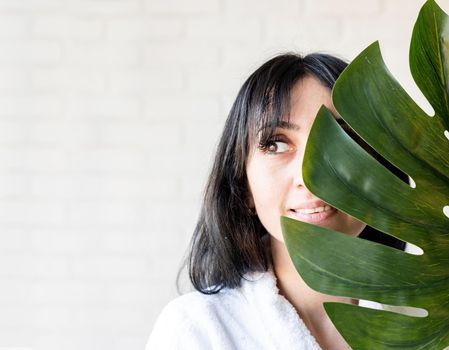 Spa Facial Mask. Spa and beauty. Happy beautiful brunette middle eastern woman wearing bath robes holding a green monstera leaf in front of her face