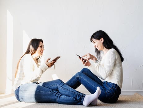 Two beautiful laughing women in jeans and white sweaters texting to friends using mobile phone