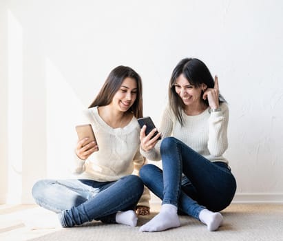 Two beautiful laughing women in jeans and white sweaters texting to friends using mobile phone