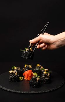 Hand with chopsticks holds a sushi. Custom sushi roll with black rice, crab meat, avocado, smoked salmon mousse, oar caviar, masago, shrimp cocktail, edible gold leaf, ginger, wasabi on black table