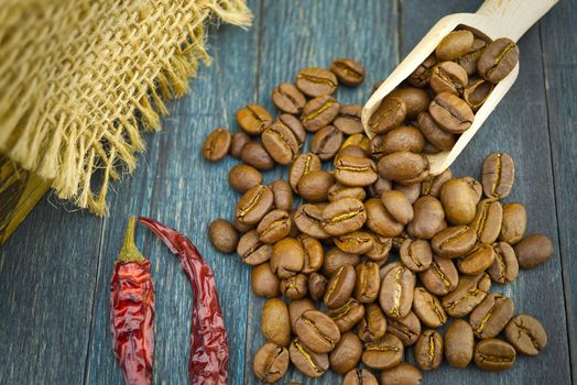 Roasted coffee beans spilling from a wooden scoop near dried red chili pepper on wooden and hessian or burlap fabric background