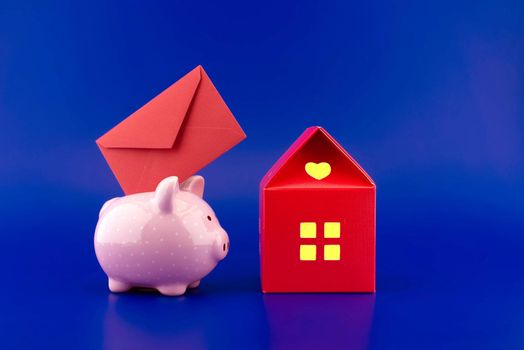 Festive red envelope being inserted into a pink piggy bank next to a small red house of paper with warm light in windows over blue background with copyspace