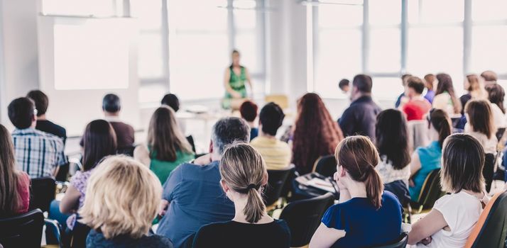 Business and entrepreneurship symposium. Female speaker giving a talk at business meeting. Audience in conference hall. Rear view of unrecognized participant in audience.