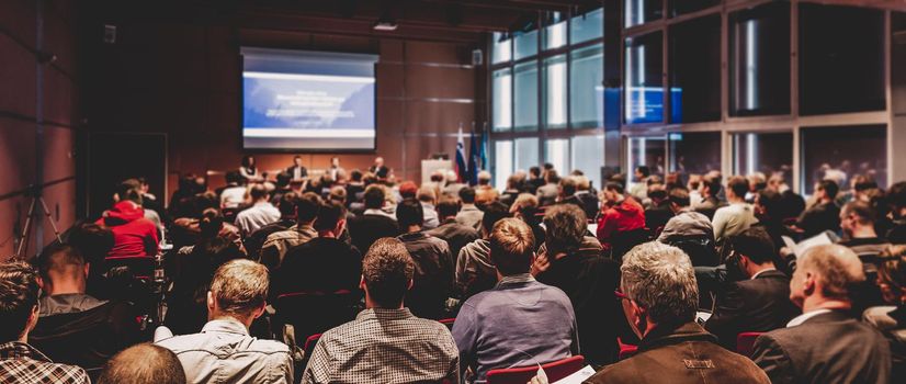 Business and entrepreneurship symposium. Speaker giving a talk at business meeting. Audience in conference hall. Rear view of unrecognized participant in audience.