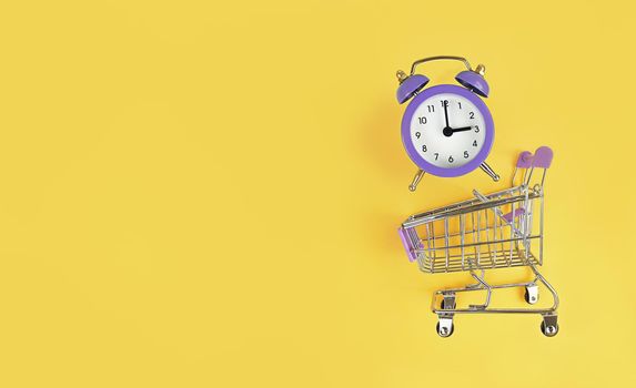 Supermarket trolley and violet alarm clock on a yellow background with copy space. Buy time, busy concept.