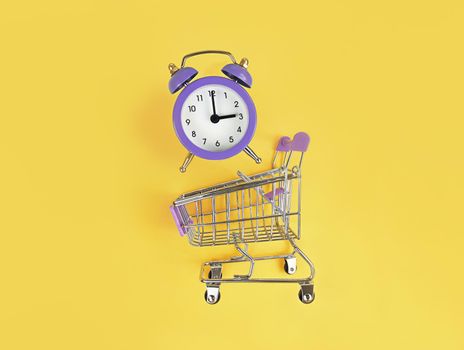 Supermarket trolley and violet alarm clock on a yellow background. Buy time, busy concept.