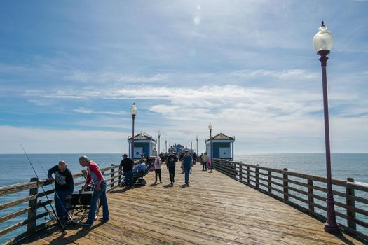 Tourist walking on the Oceanside Pier during blue summer day, Oceanside, northern San Diego County, California. Wooden pier on the western United States coastline. Famous for fisher. March 22nd, 2020