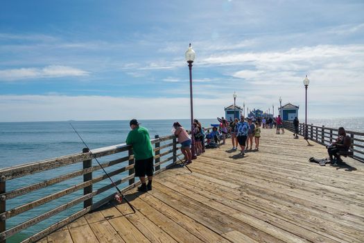 Tourist walking on the Oceanside Pier during blue summer day, Oceanside, northern San Diego County, California. Wooden pier on the western United States coastline. Famous for fisher. March 22nd, 2020