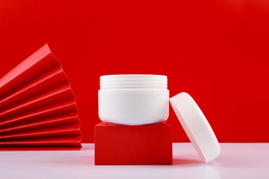 Selective focus, close up of opened white jar with cream, scrub or mask on red pedestal against red background decorated with waver. Concept of luxury skin or hair care . 