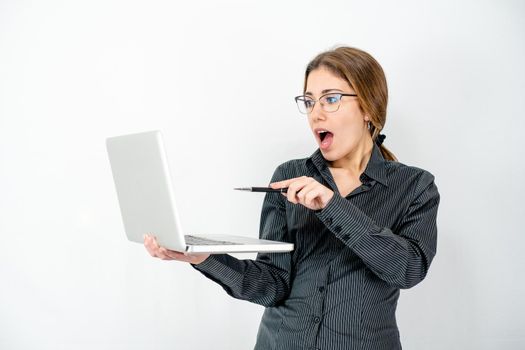 Surprised isolated beautiful young woman posing in study shot holding laptop in hand pointing with pen the screen wearing office business clothes. Caucasian female employee in wow expressions watch pc