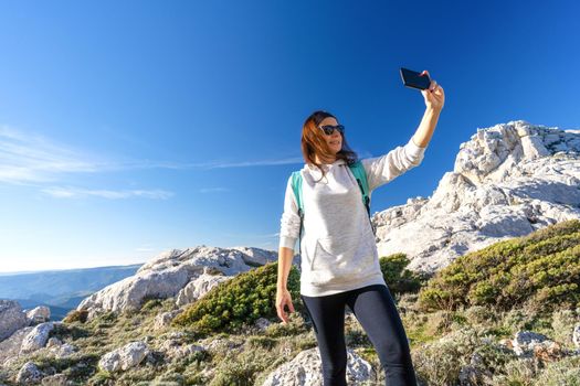 Female Caucasian hiker reaching the mountain peak take a self portrait with smartphone. Active woman with sunglasses and backpack using cellphone to share photo of her trip in nature among rocks