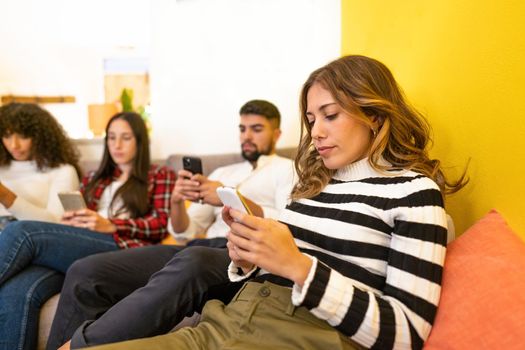 Mixed-race of technology addicted young friends serious looking at the smartphone at home on sofa without interacting with each other. New not human contacts between people in today's times