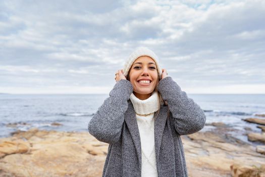 Woman tucks her wool hat on her head smiling in the cold winter outdoors at a sea resort. Beautiful young happy woman portrait in ocean coast wearing coat and sweater to stay warm in her vacation