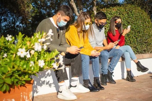 New normal social distancing for people in urban activities: group of millennial sitting on a city wall with protective mask against Coronavirus pandemic using smartphone while wait outside to enter