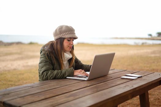New normal job activities using internet connection outdoor: young Caucasian woman sitting at a wooden table in a sea park working at laptop managing her online business. University student in nature