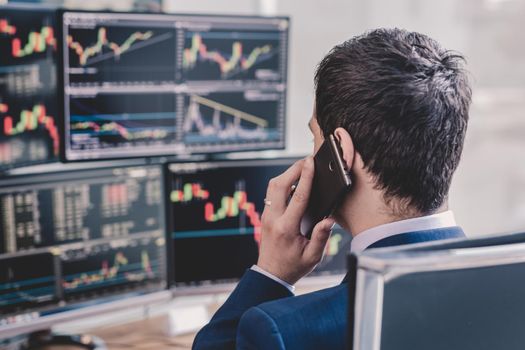 Over the shoulder view of and stock broker trading online while accepting orders by phone. Multiple computer screens ful of charts and data analyses in background.