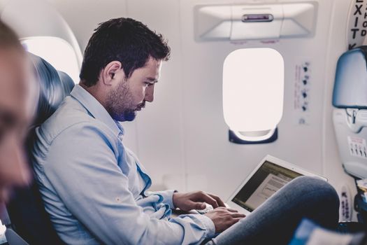 Casually dressed middle aged man working on laptop in aircraft cabin during his business travel. Shallow depth of field photo with focus on businessman eye.