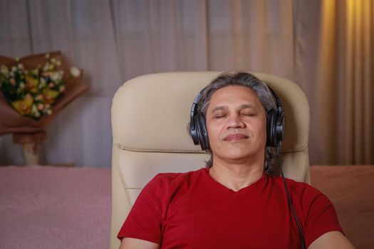 50-year-old man listens to music on headphones at home, sitting in a chair. Relaxing delight.