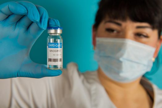 Young nurse in a medical mask holding vial of the Covid-19 coronavirus vaccine on a blue background. Close-up. Selective focus.